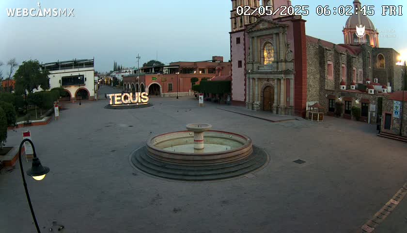 A town square with a fountain at its center is shown under a twilight sky, with a large church visible in the background.