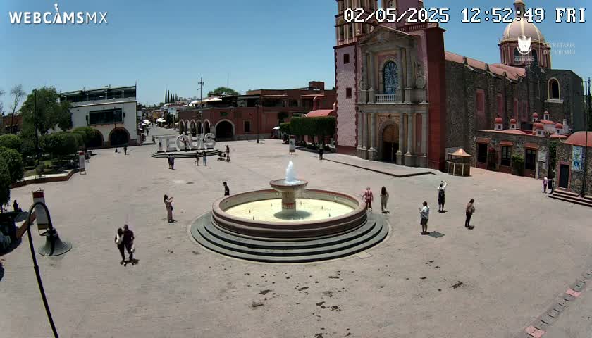 A town square with a fountain, several people, and a large church under a clear, sunny sky.