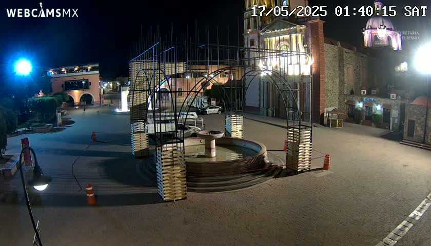 A nighttime view of a town square with a fountain, buildings, and a metal structure, under clear skies.