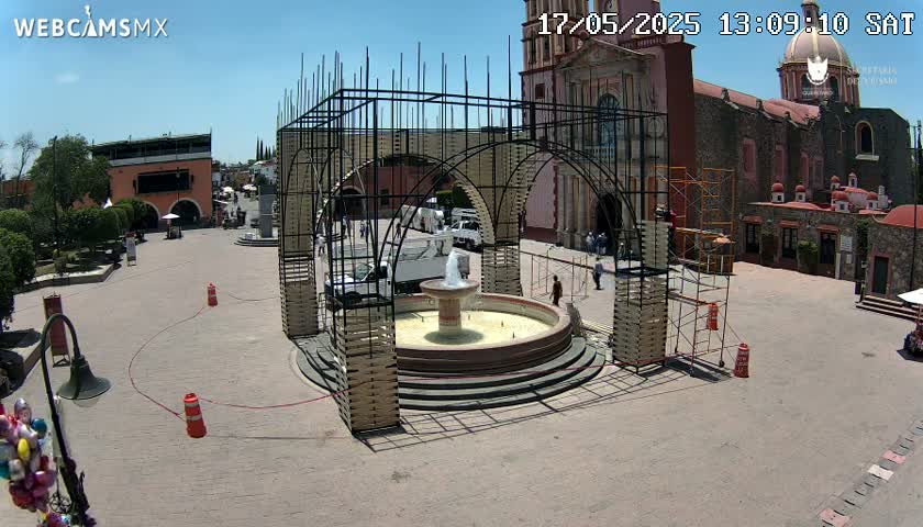 A town square under a clear blue sky features a fountain surrounded by a metal structure, with a church in the background.