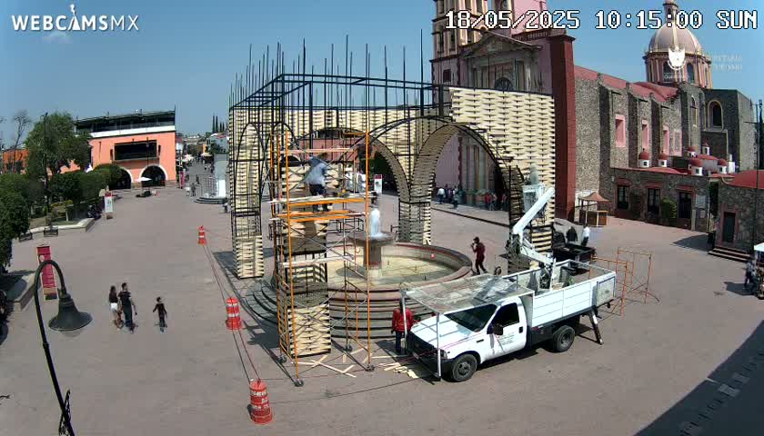 Under a clear, sunny sky, construction workers erect a large archway structure in a town square next to a church, with a fountain in the center.