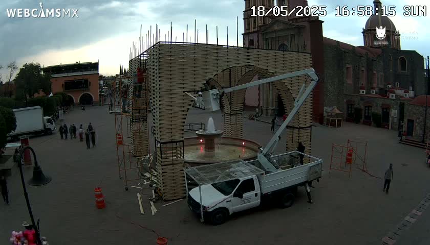 A large, arched structure is under construction in a town square near a stone church, under an overcast sky; a white truck with a lift is nearby.