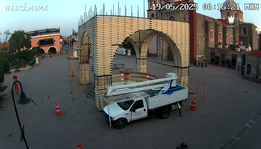 A large, light-brown arched structure is under construction in a town square next to a church, with a bucket truck parked nearby under a clear morning sky.