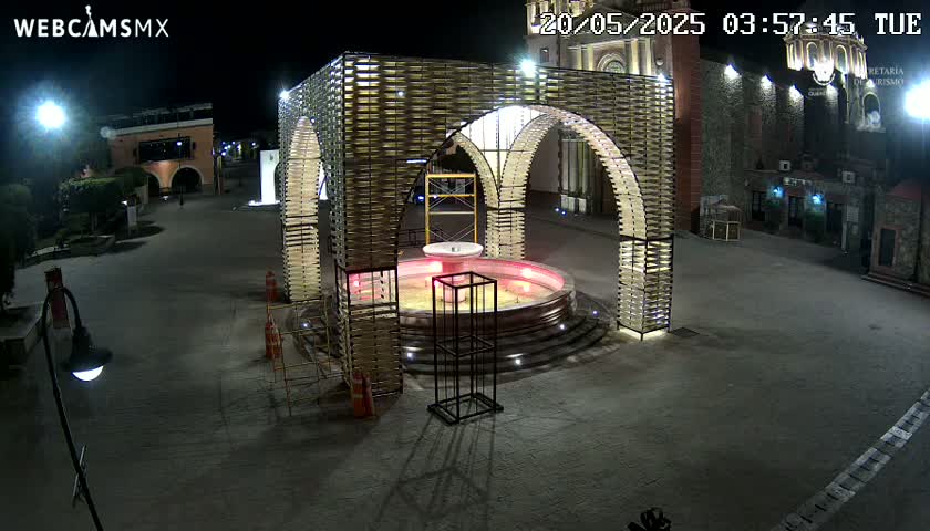 A brightly lit fountain sits at the center of a plaza at night, surrounded by a modern, arched structure.