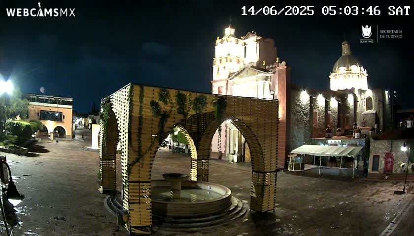 A nighttime view of a town square with a fountain under a latticed structure, illuminated buildings including a church, and a mostly clear sky.