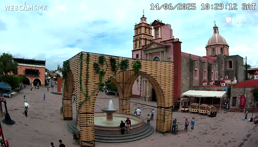 A town square with a large, ornate fountain, a wooden archway structure decorated with greenery, and a church in the background under partly cloudy skies.