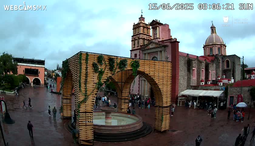 A town square with a large, ornate wooden structure surrounding a fountain, situated in front of a church under an overcast sky.