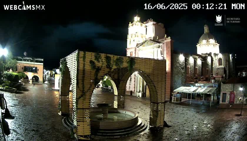 A town square with a large, ornate wooden structure surrounding a fountain, situated in front of a church under an overcast sky.