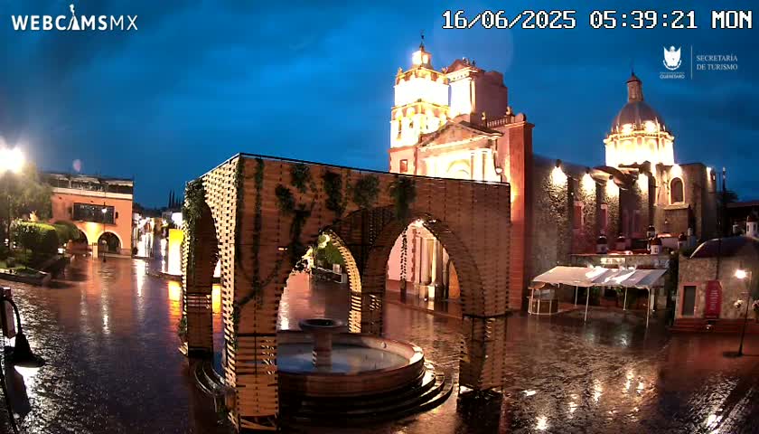 A rain-soaked town square at night, featuring a large illuminated church and a modern wooden structure surrounding a fountain.