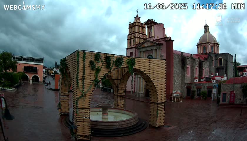 A town square with a large, ornate fountain is situated in front of a church under an overcast sky.