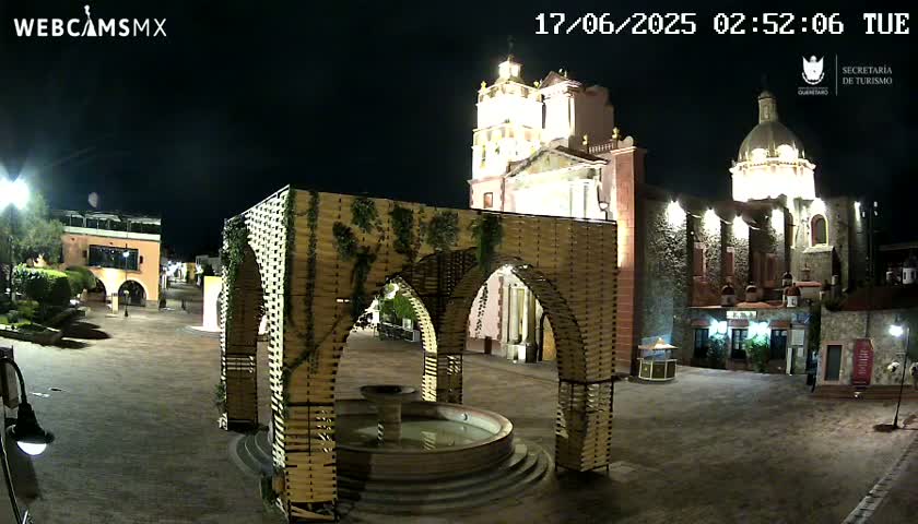 A nighttime view of a town square with a fountain under a wooden archway structure, illuminated buildings, and a church in the background.