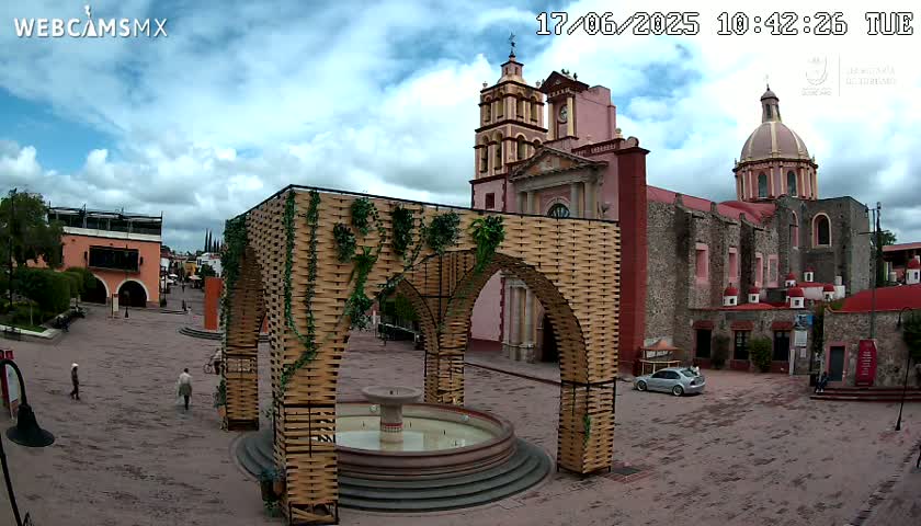 A town square with a large, ornate archway structure and a fountain, situated in front of a pink and grey church under a partly cloudy sky.