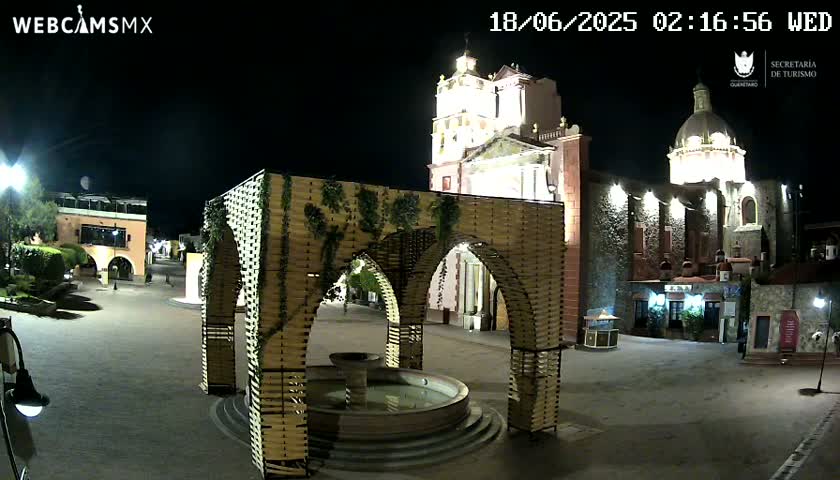 A nighttime view of a town square with a fountain under a decorative archway, adjacent to a large, illuminated church.