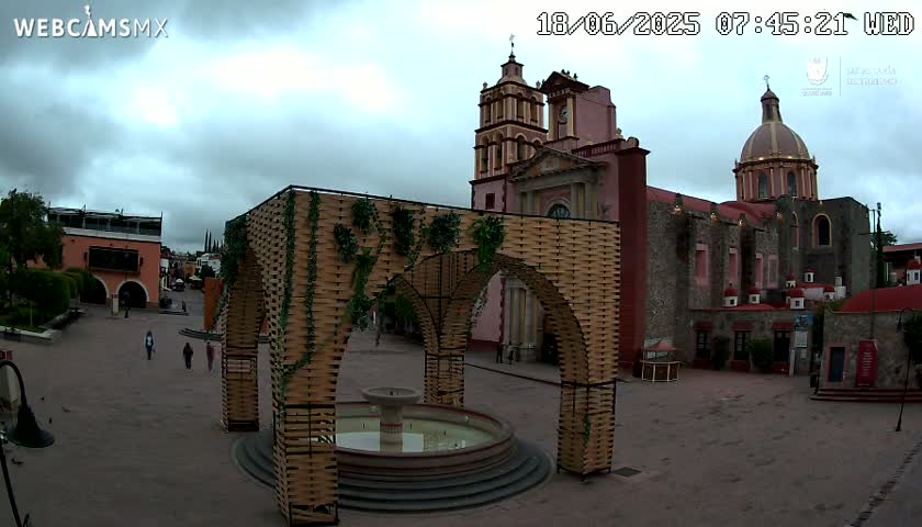 A town square with a large, ornate fountain under a woven archway, situated in front of a church under an overcast sky.