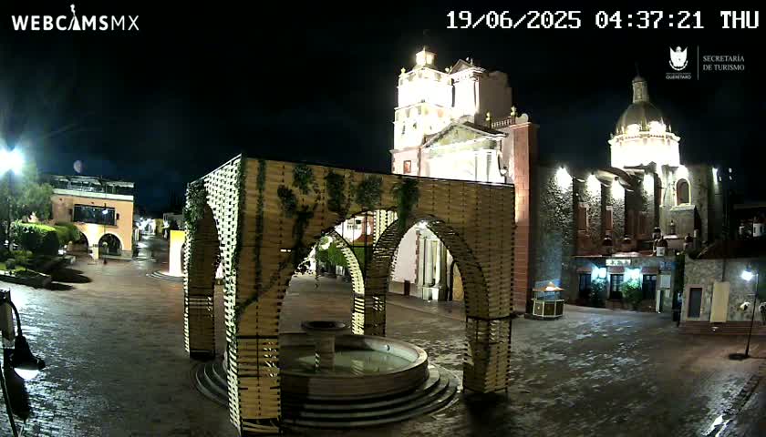 A nighttime view of a town square with a fountain, a modern archway structure, and illuminated colonial-style buildings under a clear sky.