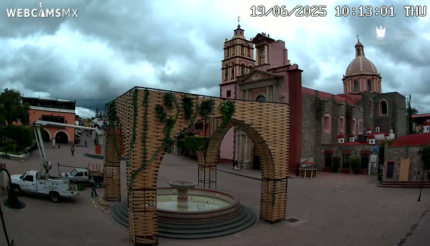 A town square under a cloudy sky features a large, ornate archway structure near a fountain, with a church in the background.