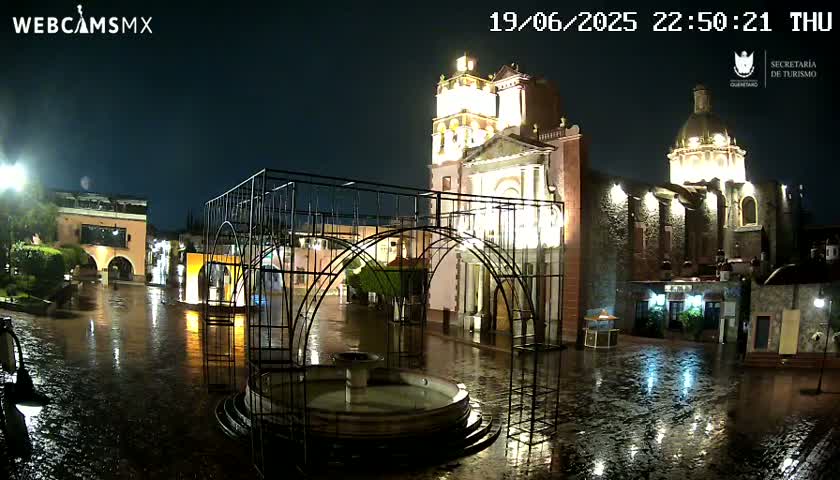 A rain-slicked plaza at night, illuminated by lights, features a large fountain and two illuminated churches.