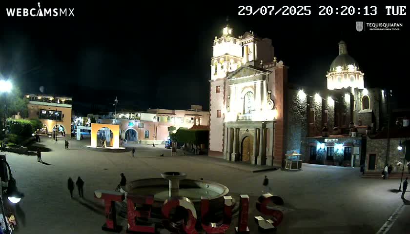 A nighttime view of a town square with a fountain, a large illuminated church, and surrounding buildings under a clear, dark sky.