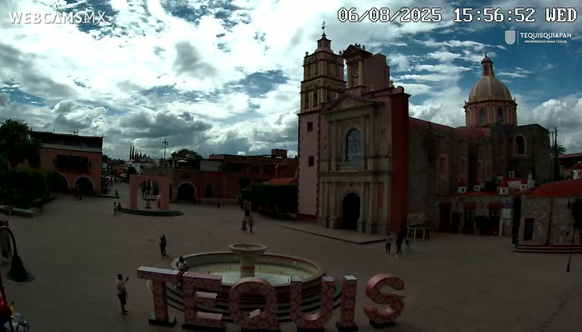 A town square with a large pink church and a fountain, under a partly cloudy sky.