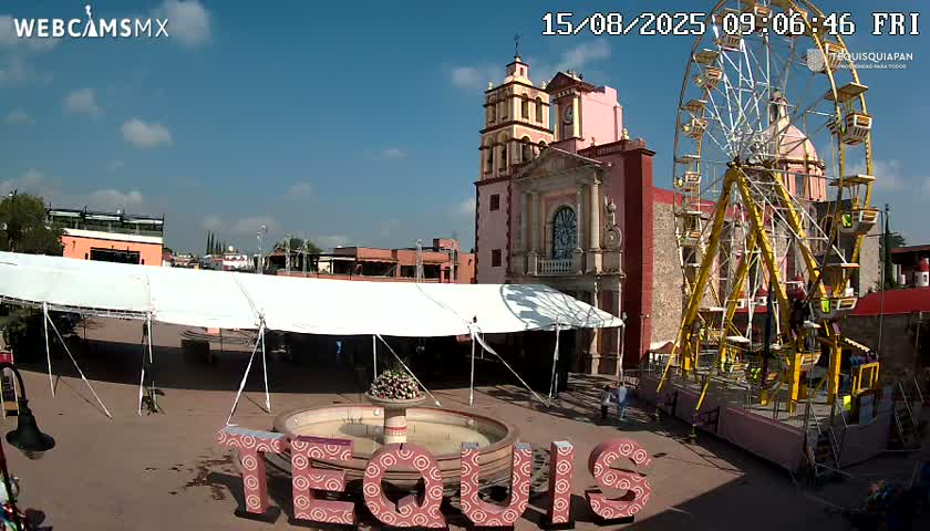 A town square with a large pink church and a fountain, under a partly cloudy sky.