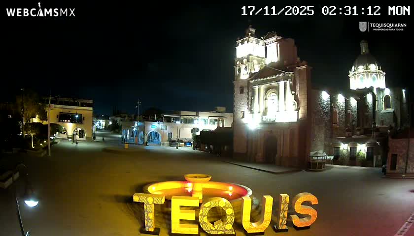 A brightly lit town square is captured at night under a clear sky, featuring a grand church with a tall tower and a dome, surrounded by buildings with illuminated arcades, and a decorative glowing fountain with large letters in the foreground.