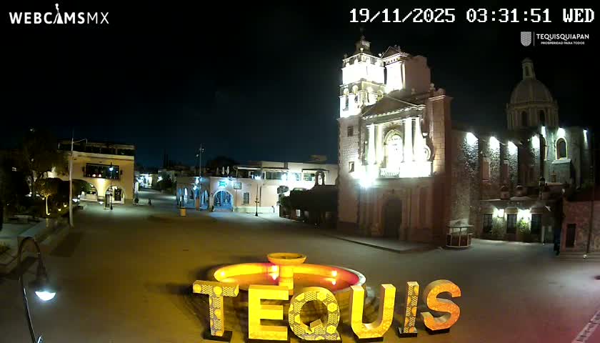 This nighttime image shows a brightly lit central plaza featuring a grand church with a bell tower and dome, surrounded by other traditional buildings, all under a clear, dark sky.