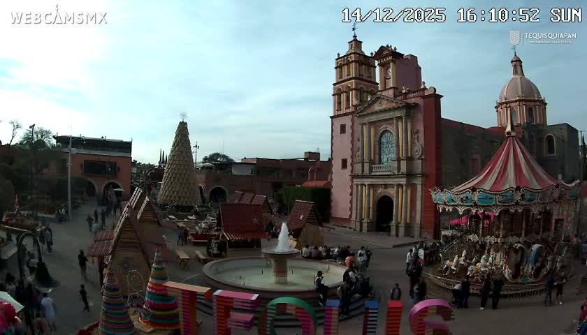 A brightly illuminated historic church with a prominent bell tower and dome overlooks a quiet, empty town square surrounded by warmly lit colonial-style buildings at night, under a clear sky.