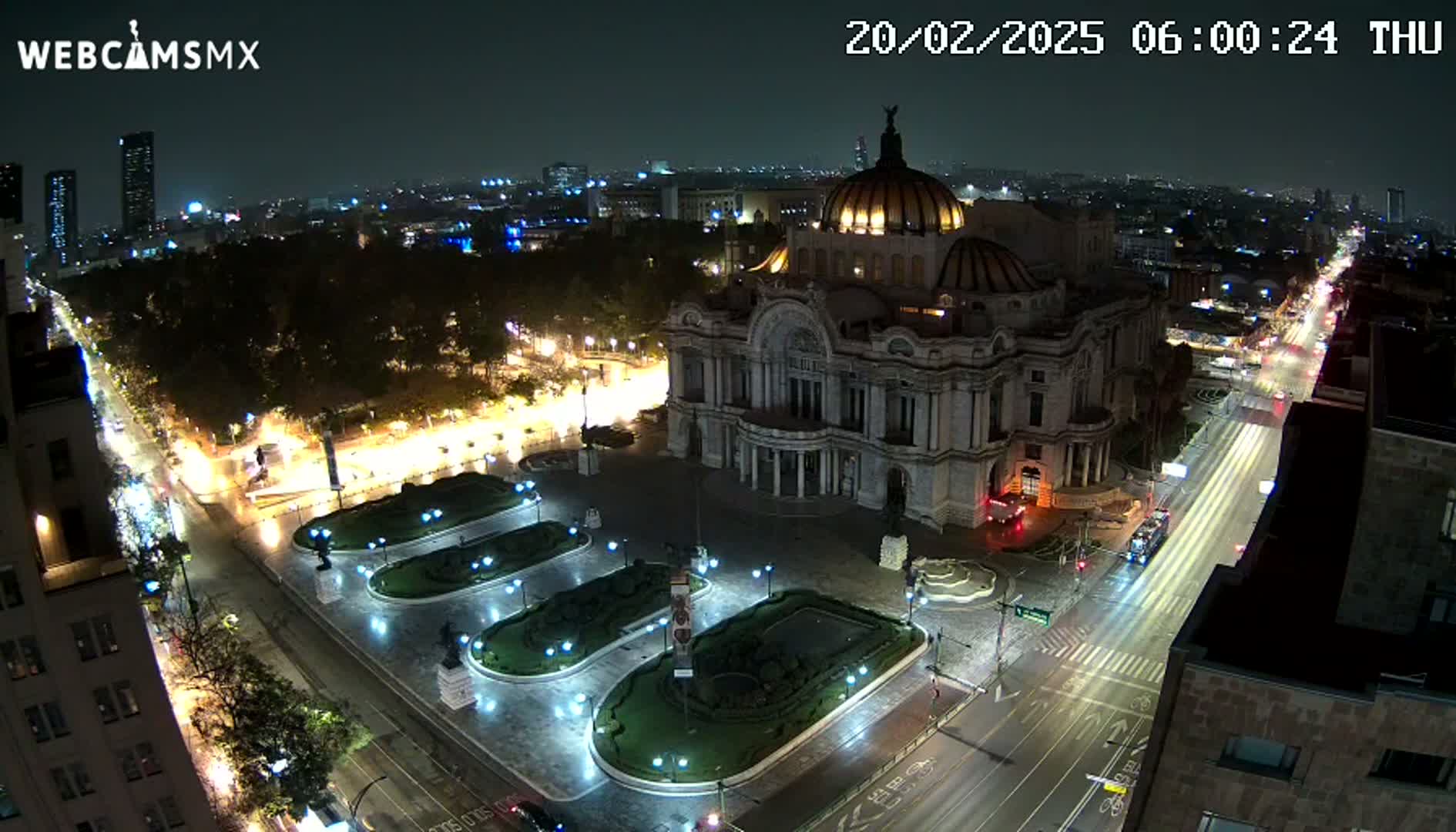 A nighttime, high-angle view shows a large, ornate building, possibly a museum or palace, situated in a city square with wet streets and surrounding illuminated landscaping, all under a dark sky.