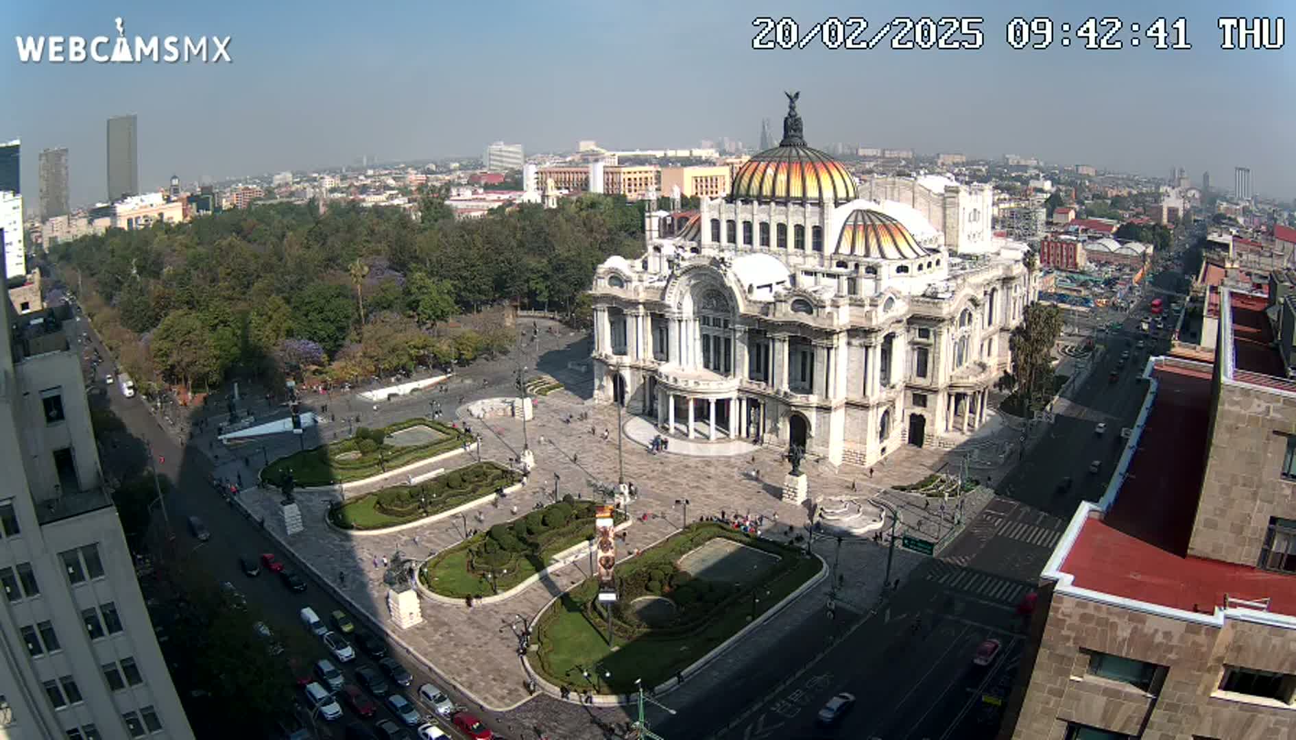 A large, ornate palace-like building sits in a plaza with manicured gardens, surrounded by city streets and buildings on a sunny day.