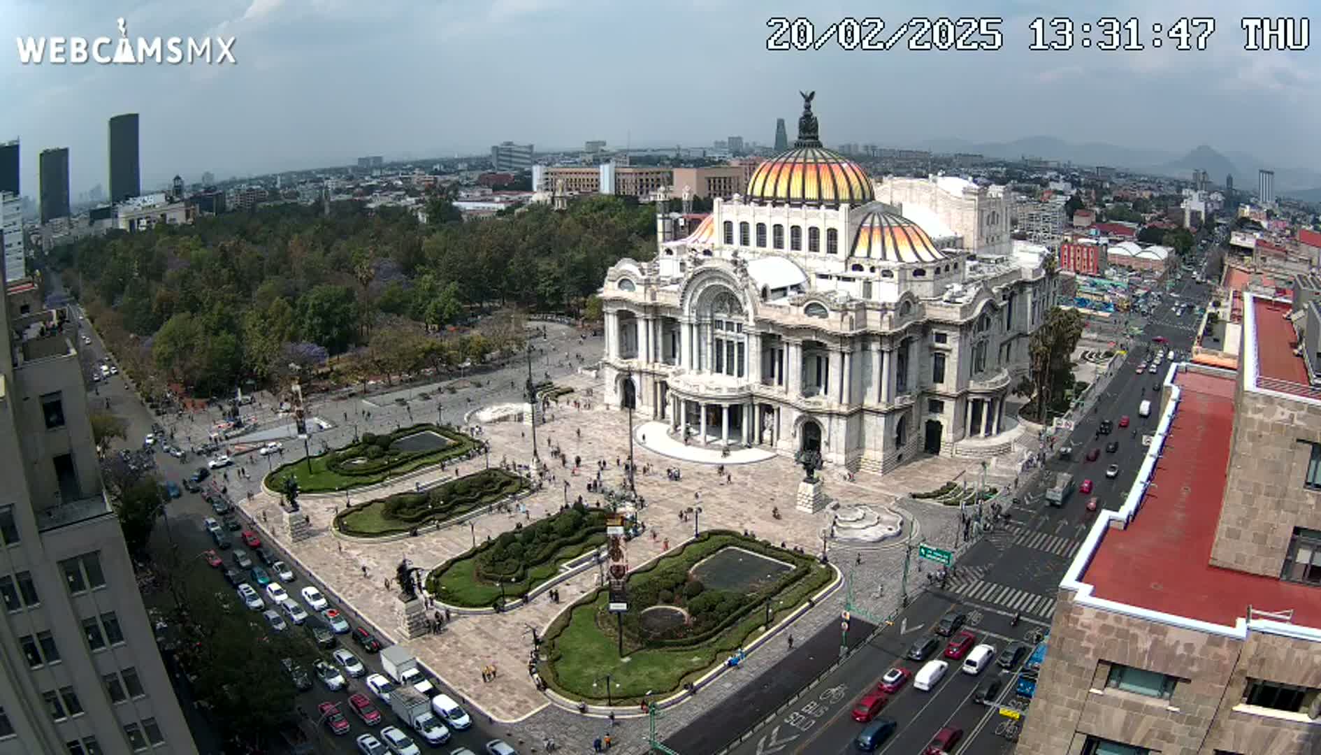 An aerial view shows a large, ornate building surrounded by a plaza with landscaped gardens and a city street with cars under a partly cloudy sky.