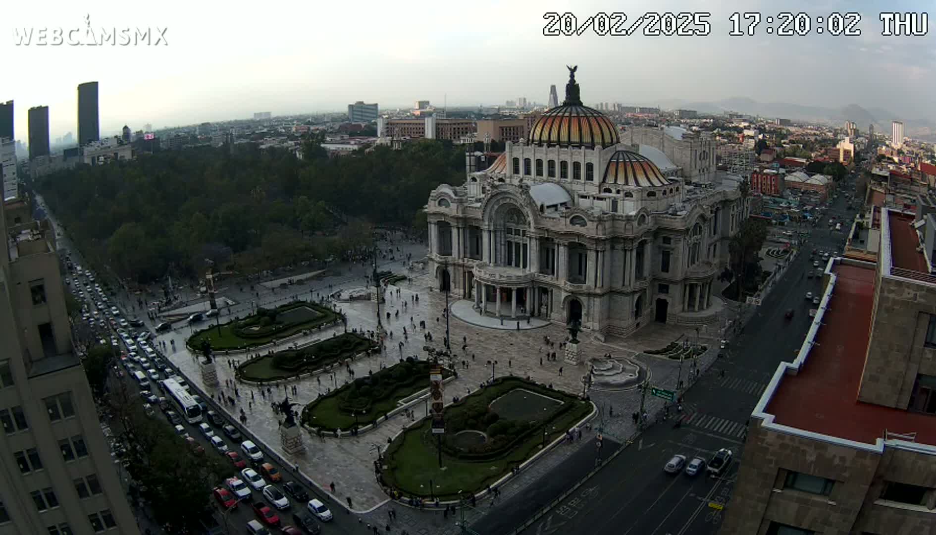 An aerial view shows the Palacio de Bellas Artes in Mexico City on an overcast day, with traffic moving along a nearby street and people walking in the plaza surrounding the building.