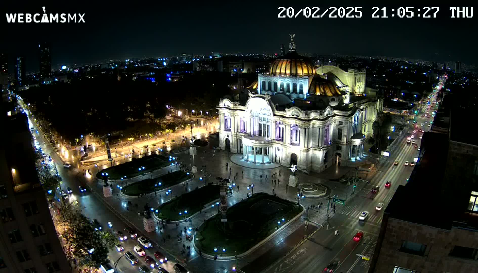 A nighttime, aerial view of a large, ornate building, brightly lit, surrounded by city streets with moving cars and a park-like area.