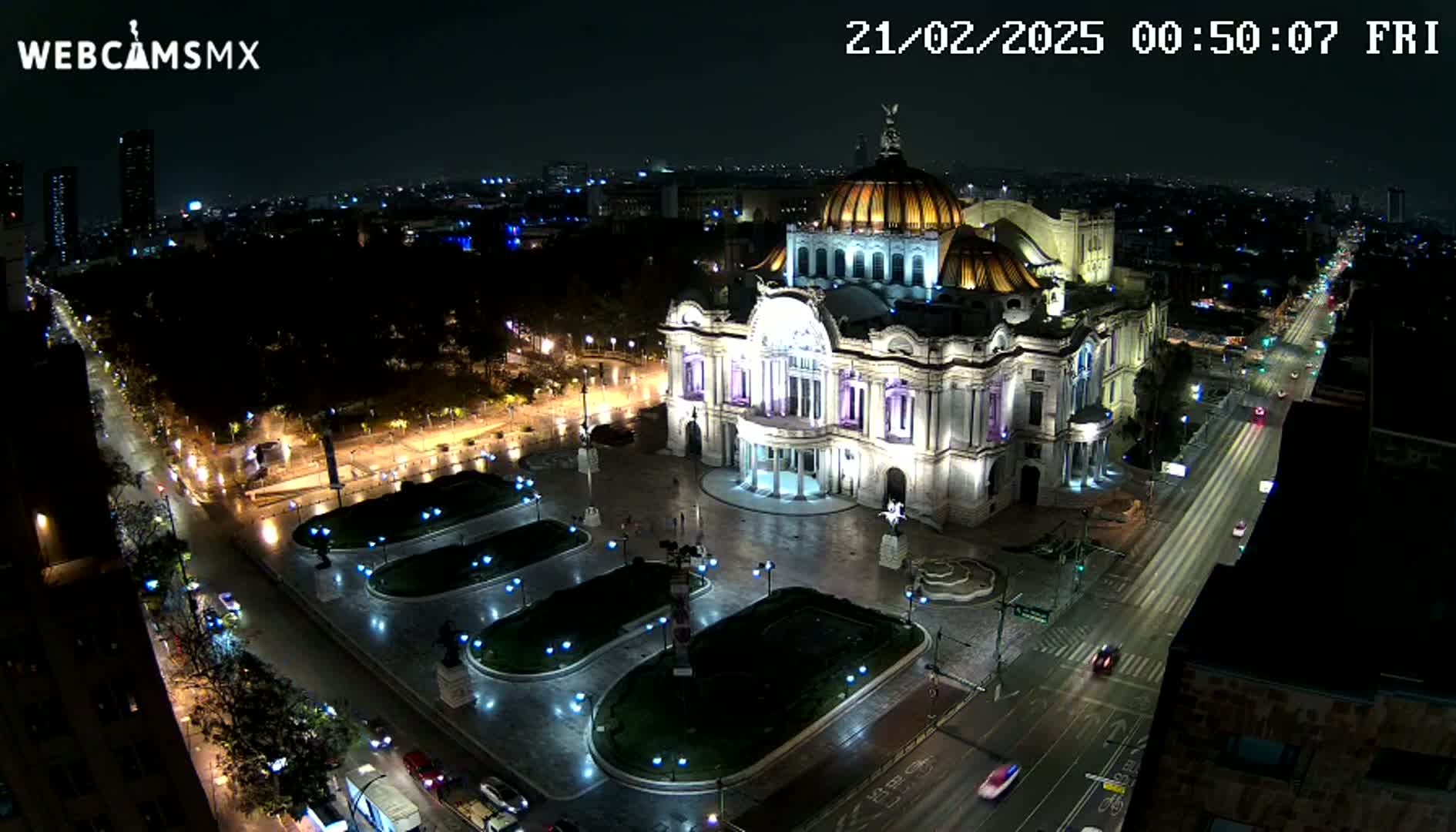 A brightly lit Palacio de Bellas Artes stands in a plaza at night, with a city skyline and streets visible in the background under a dark sky.