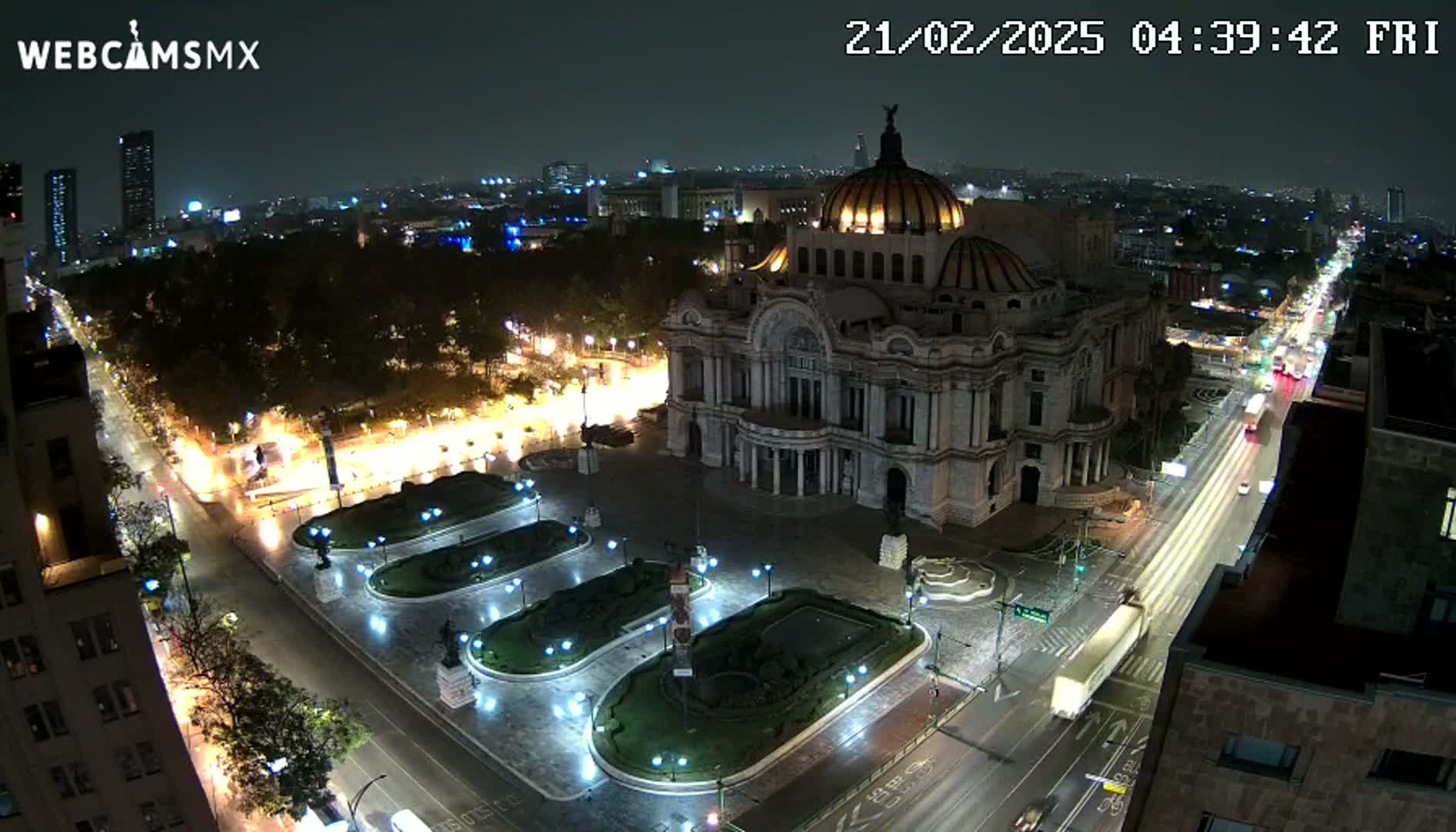 A nighttime, aerial view of a large building, possibly a museum or palace, situated in a city square with wet pavement, surrounded by illuminated streets and park-like areas.