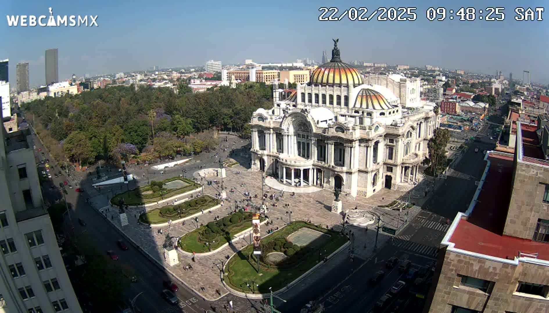 A large, ornate building with a dome sits in a plaza surrounded by trees, landscaping, and streets with cars, under a clear, sunny sky.