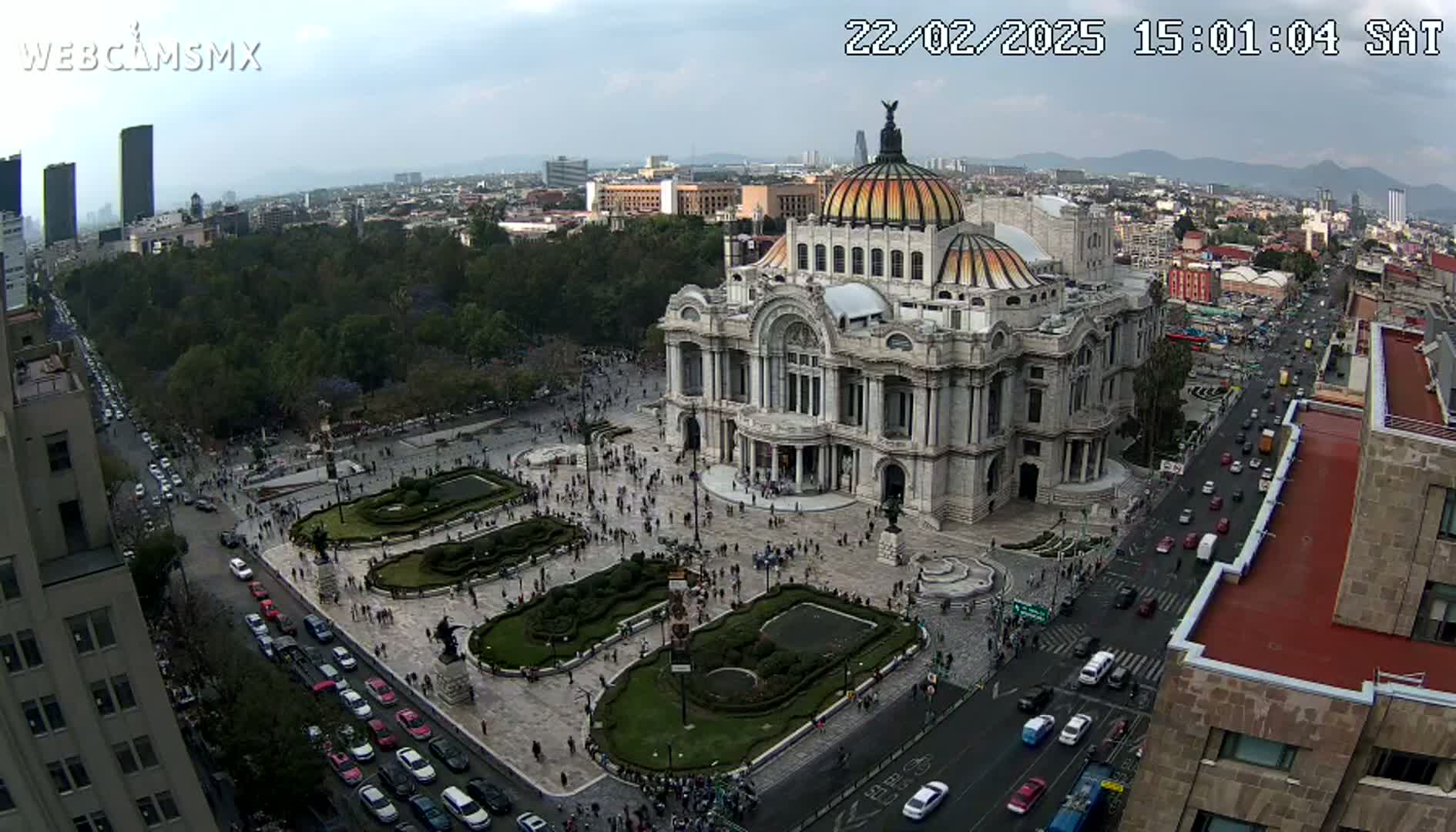An aerial view of a large, ornate building surrounded by a plaza with landscaping and people, situated in a city under a mostly cloudy sky.