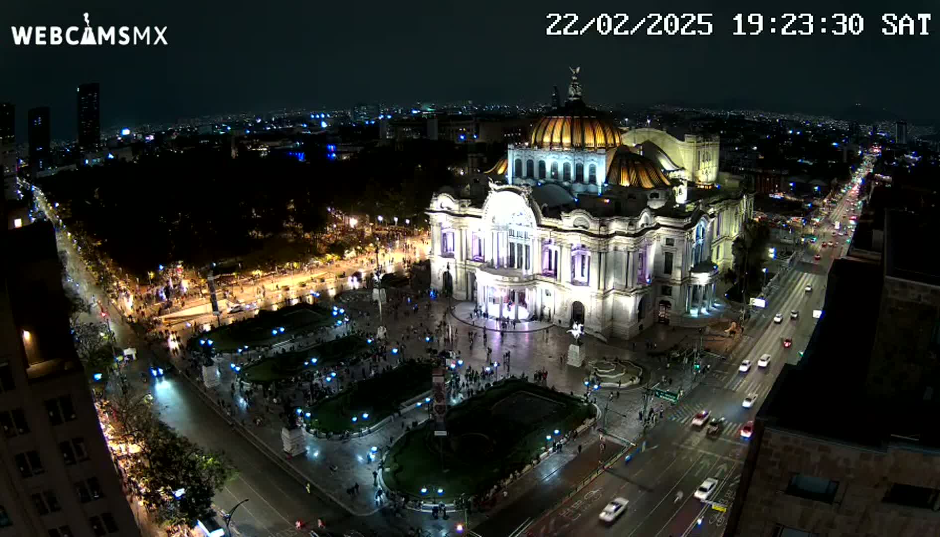 An illuminated Palacio de Bellas Artes sits in a city square at night, surrounded by streets with cars and pedestrians under a clear sky.
