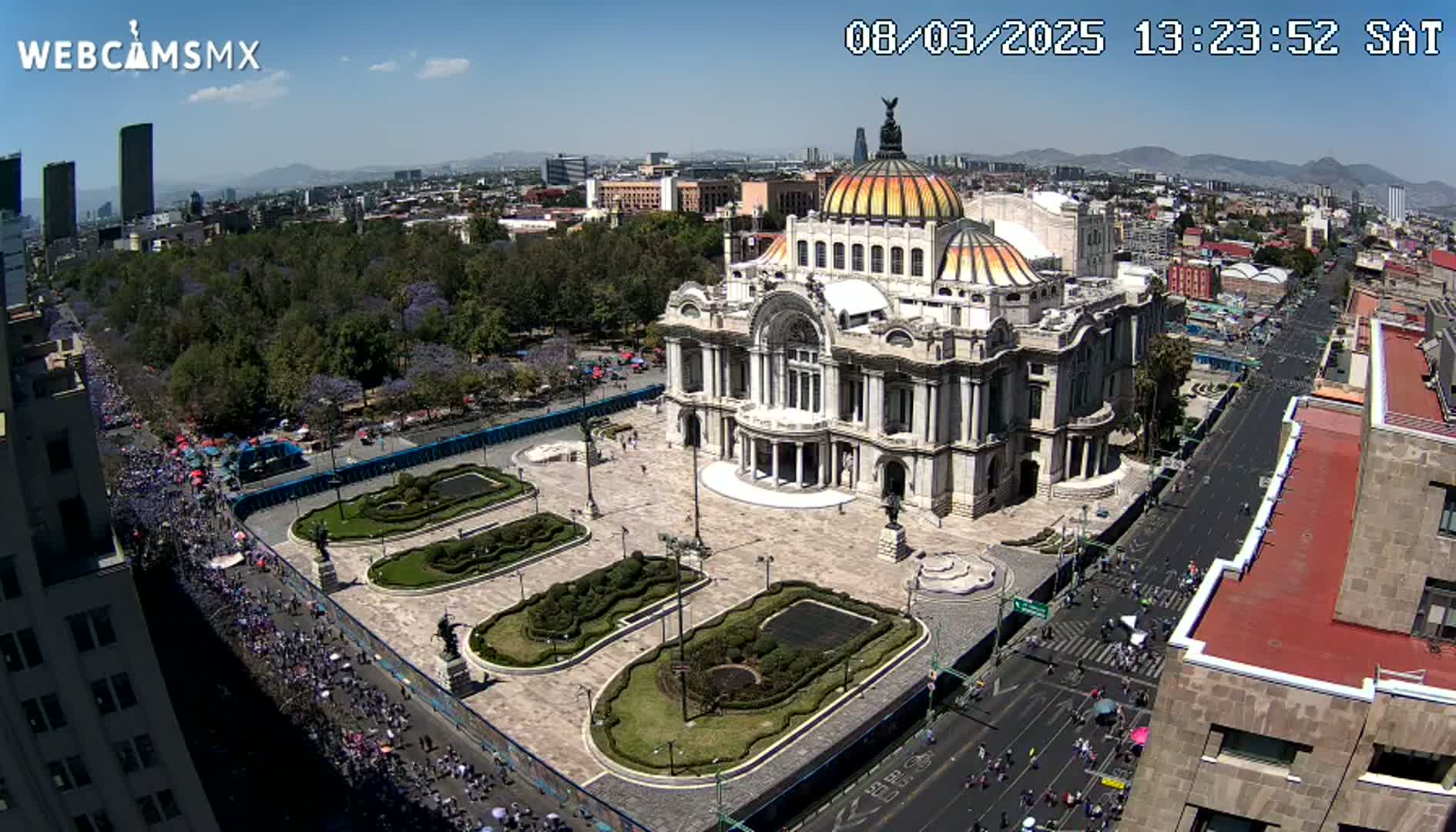 An aerial view on a sunny day shows a large, ornate building surrounded by a plaza with landscaped gardens, and a street filled with people.
