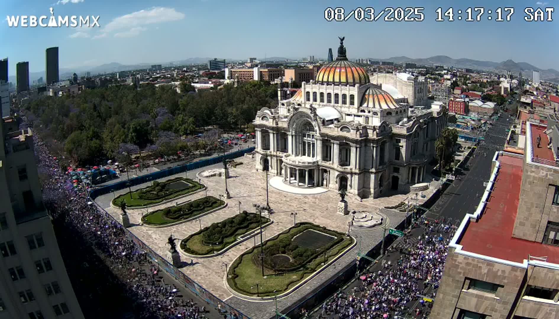 A large crowd marches along a city street past a grand, ornate building with a dome under a sunny sky.