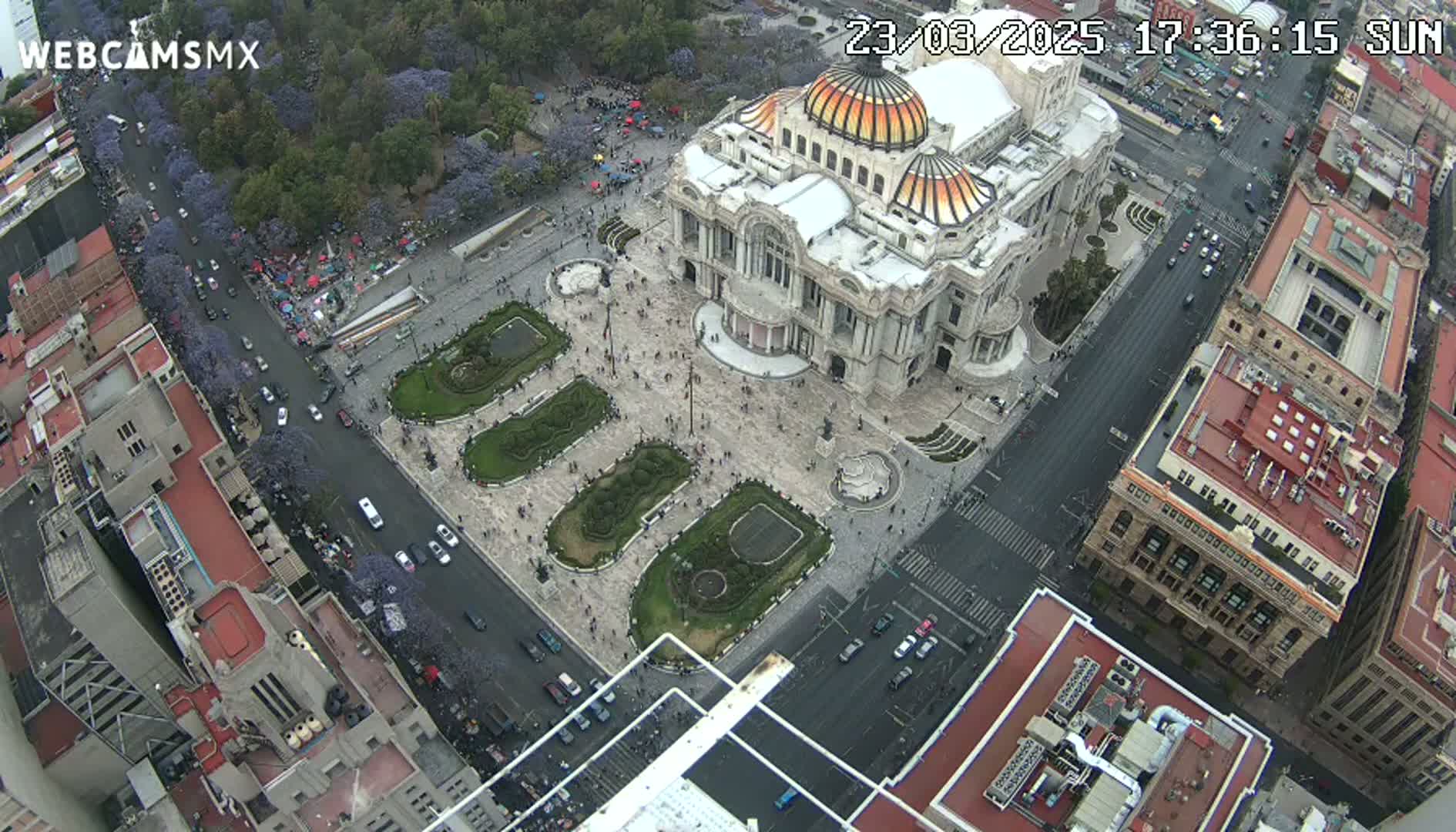 An aerial view shows a large, ornate building situated in a plaza with landscaped gardens, surrounded by city streets and buildings on a partly cloudy day.