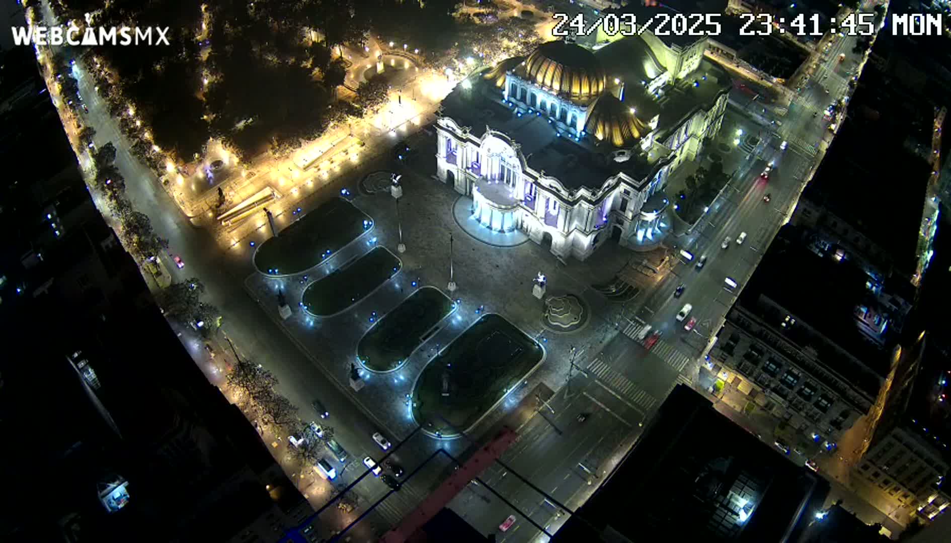 An illuminated Palacio de Bellas Artes sits in a plaza at night, surrounded by city streets with cars and buildings.