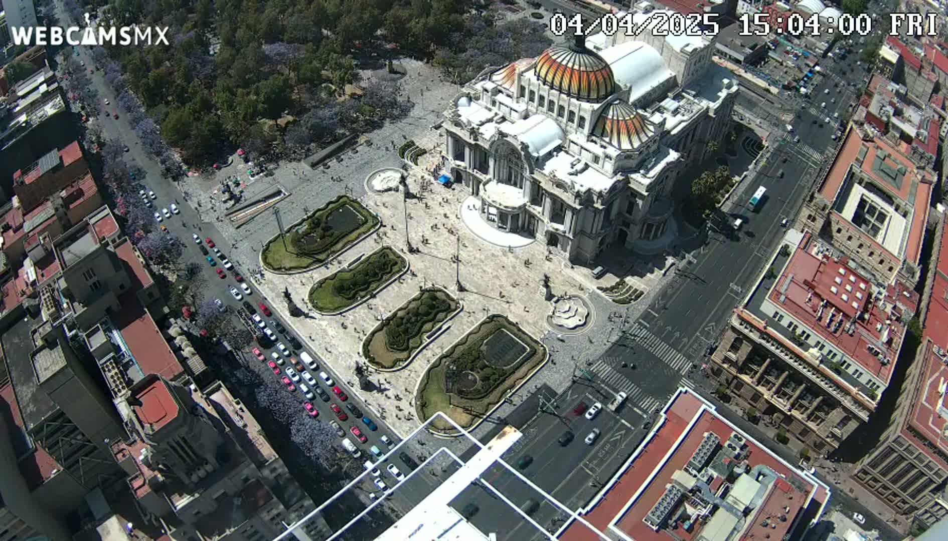 An aerial view shows a large, ornate building situated in a plaza with landscaped gardens, surrounded by city streets and buildings on a sunny day.