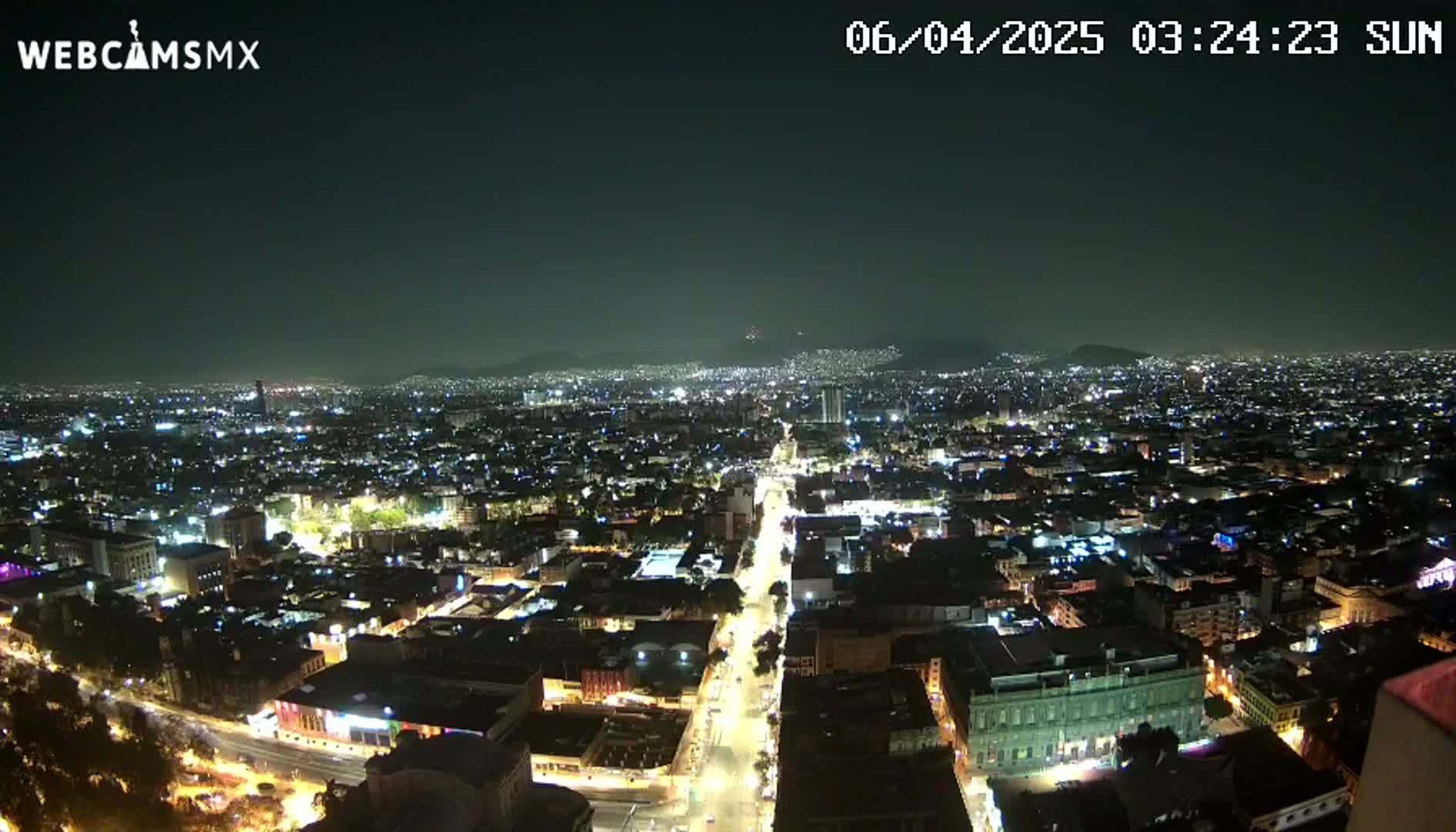 A nighttime, high-angle view of a sprawling city illuminated by countless lights, with distant mountains visible under a clear, dark sky.