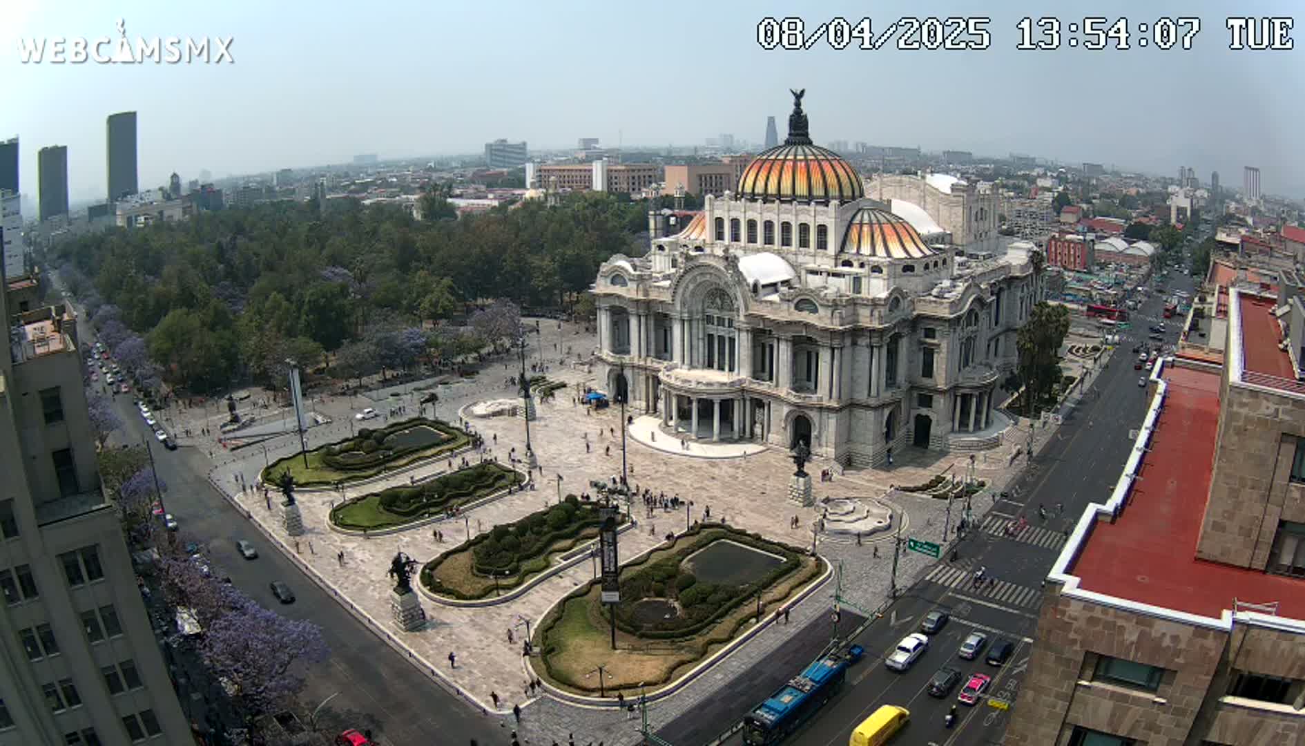 An aerial view shows a large, ornate building in a city square surrounded by landscaping and a street with cars, on a hazy, sunny day.