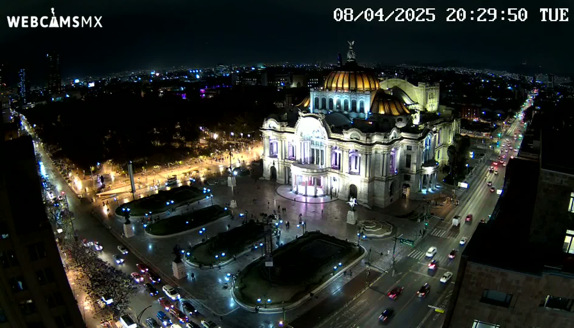An illuminated Palacio de Bellas Artes sits in a plaza at night, surrounded by city streets with moving vehicles under a clear sky.