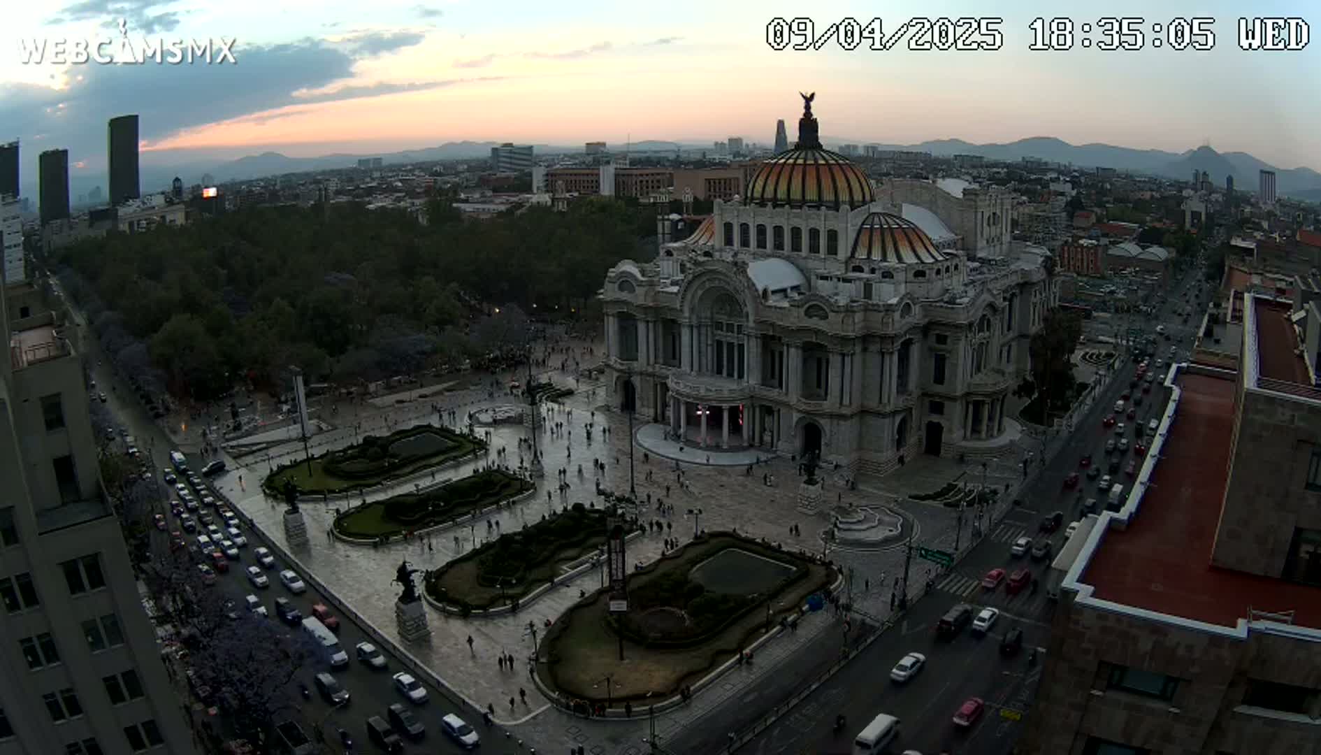 An aerial view shows Palacio de Bellas Artes in Mexico City at dusk, with traffic on the surrounding streets and people in the plaza.