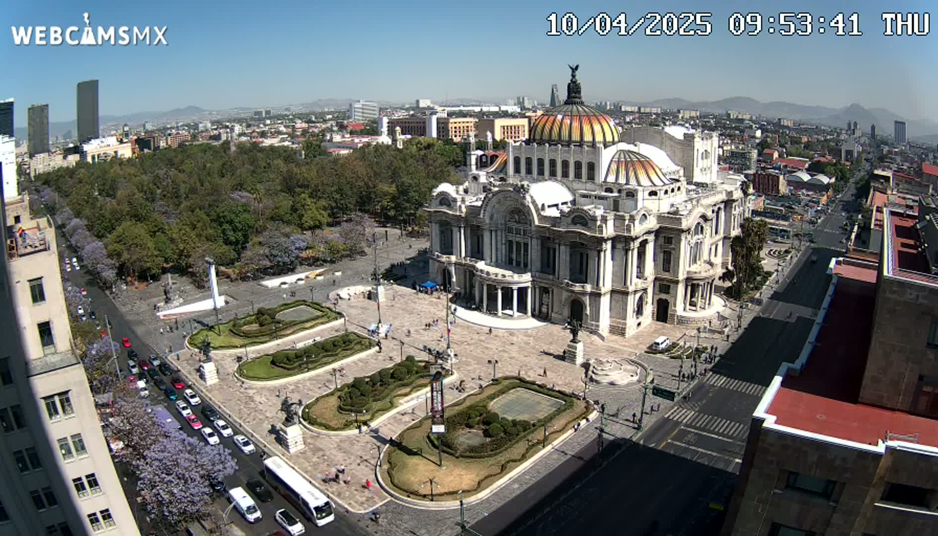 An aerial view on a sunny day shows a large, ornate building surrounded by a plaza with landscaped gardens and a city street with cars and pedestrians.