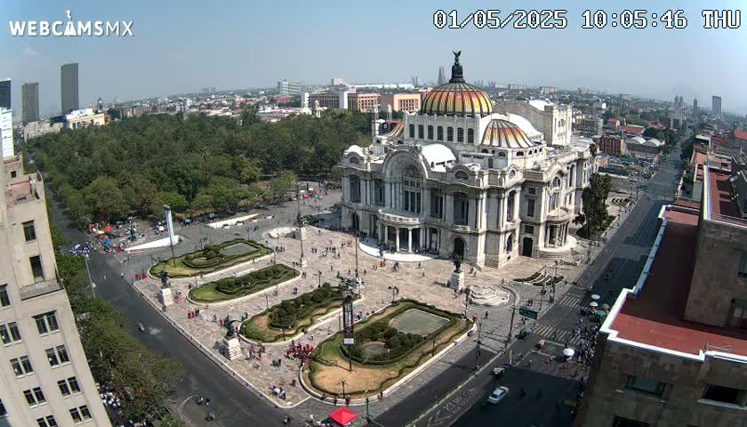 An aerial view shows a large, ornate building in a city square on a sunny day, surrounded by trees, landscaping, and streets with sparse traffic.