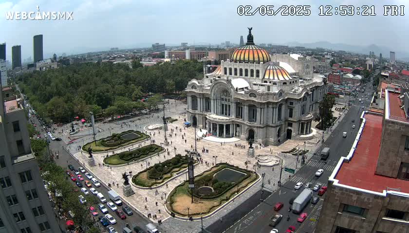 An aerial view shows a large, ornate building surrounded by a plaza with landscaping, streets with cars, and a city in the background under a mostly sunny sky.