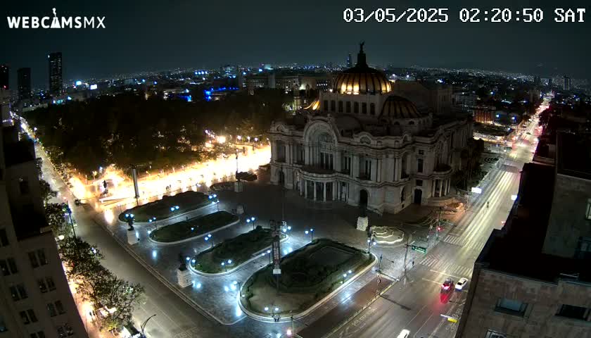 A nighttime aerial view of a large, ornate building surrounded by a plaza and city streets with sparse traffic under a clear sky.